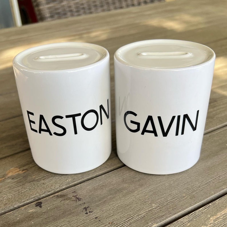 May include: Two white ceramic money banks with a slot on top. The word "EASTON" is printed in black on the first bank, and "GAVIN" is printed on the second. The banks are cylindrical and sit on a wooden surface.