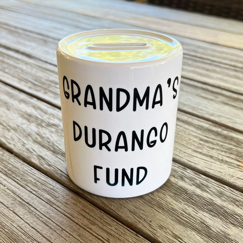 May include: White ceramic money box with a slot on top. The words "GRANDMA'S DURANGO FUND" are printed in black. The money box is cylindrical and rests on a wooden surface.