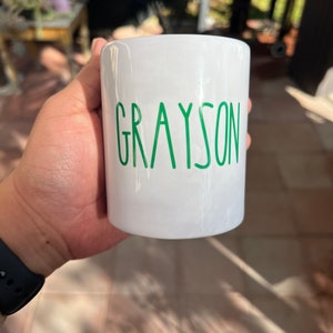 May include: A white ceramic mug with the name "GRAYSON" printed in green, sans-serif font. The mug is cylindrical and appears to be held by a person. The background is blurred, suggesting an outdoor setting.