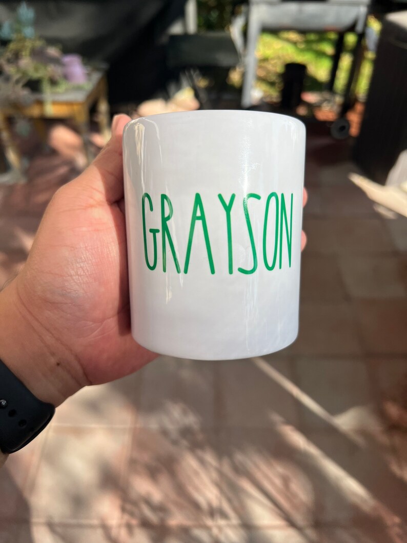 May include: A white ceramic mug with the name "GRAYSON" printed in green, sans-serif font. The mug is cylindrical and appears to be held by a person. The background is blurred, suggesting an outdoor setting.