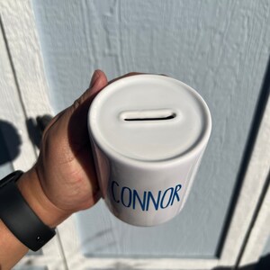 May include: White ceramic coin bank with the name "CONNOR" in blue script. The bank has a slot on top for inserting coins. The bank is cylindrical in shape.