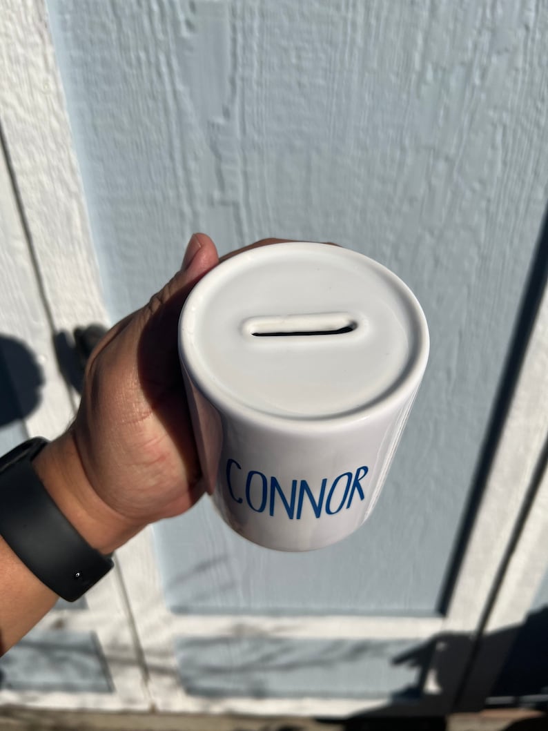 May include: White ceramic coin bank with the name "CONNOR" in blue script. The bank has a slot on top for inserting coins. The bank is cylindrical in shape.