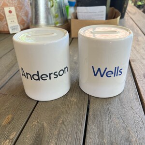 May include: Two white ceramic money banks with a slot on top. One bank has the name "Anderson" printed in black, and the other has "Wells" printed in blue. Both banks are cylindrical and sit on a wooden surface.