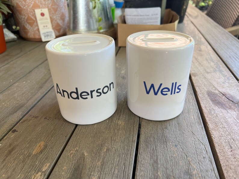 May include: Two white ceramic money banks with a slot on top. One bank has the name "Anderson" printed in black, and the other has "Wells" printed in blue. Both banks are cylindrical and sit on a wooden surface.