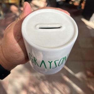 May include: White ceramic money box with a slot for coins. The name "GRAYSON" is written in green on the side. The money box is cylindrical in shape.