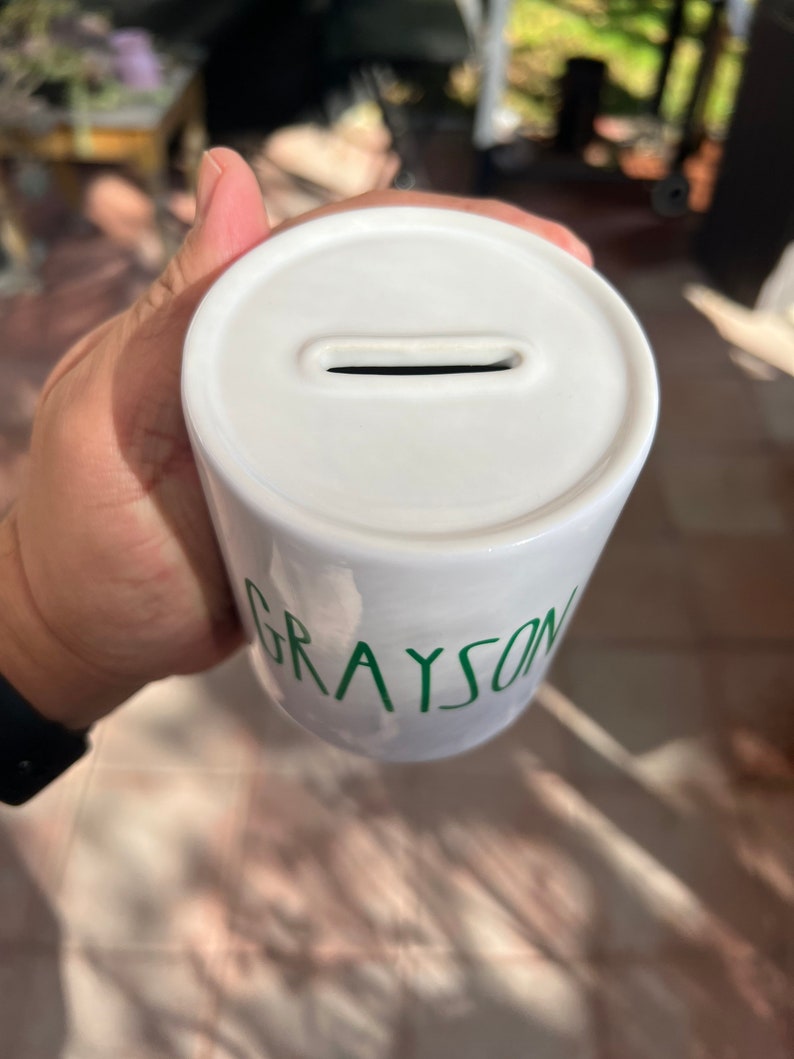 May include: White ceramic money box with a slot for coins. The name "GRAYSON" is written in green on the side. The money box is cylindrical in shape.