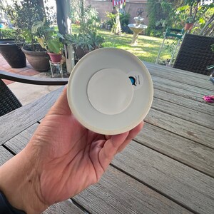 May include: A white ceramic object with a circular white sticker on the base. The object is held in a hand, with a blurred outdoor background of a garden and patio furniture.