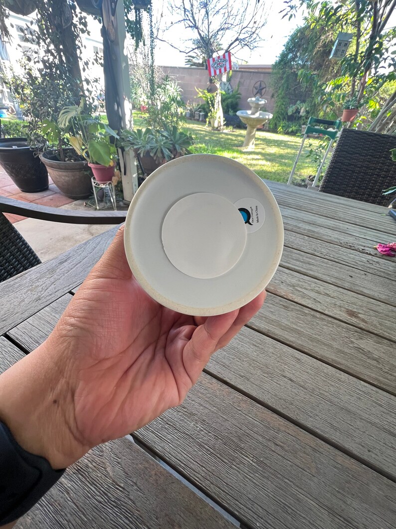May include: A white ceramic object with a circular white sticker on the base. The object is held in a hand, with a blurred outdoor background of a garden and patio furniture.
