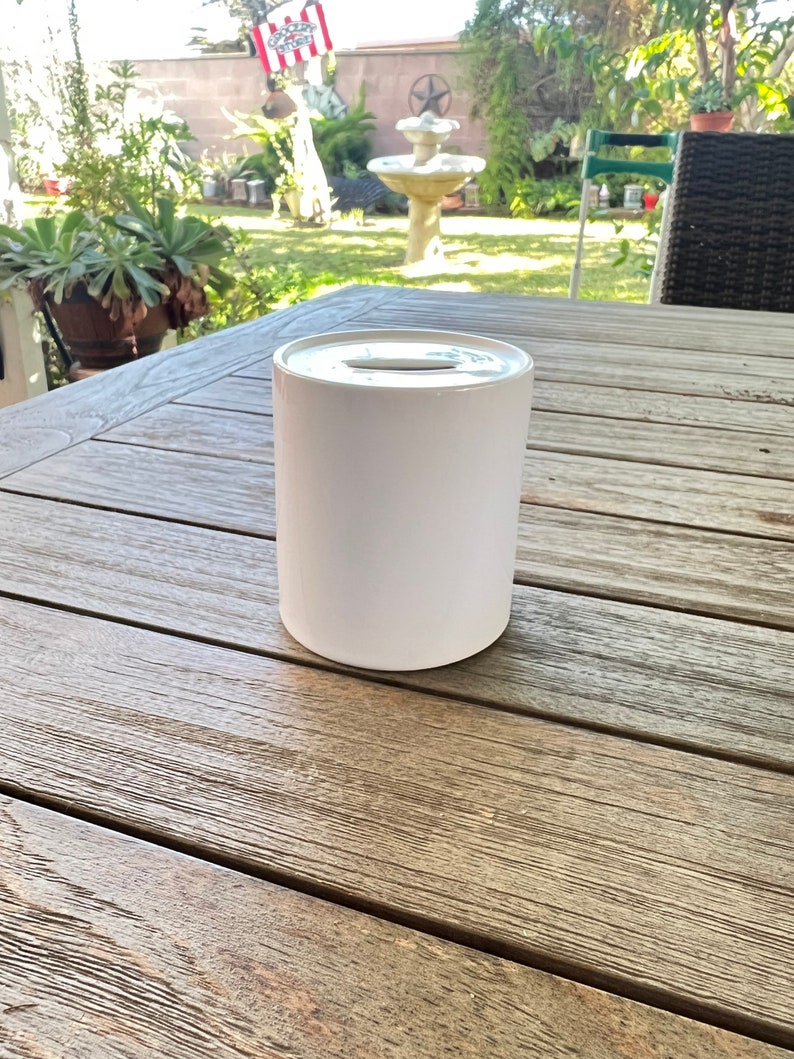May include: A white cylindrical container with a slot on top, possibly a donation box. It sits on a weathered wooden table. The background is blurred, showing a garden with plants and a fountain.
