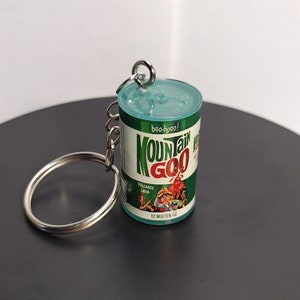 Puede incluir: Un llavero verde y blanco con forma de lata de Mountain Goo, una marca ficticia de goo. El llavero tiene un llavero plateado adjunto. La lata tiene una imagen de un hombre y una mujer de dibujos animados. El texto en la lata dice "Mountain Goo" y "Volcanic Lava".