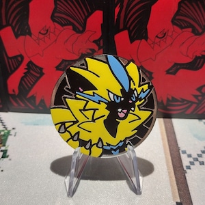 Op de afbeelding: Een gele en blauwe Pokemon-munt met een gedetailleerde illustratie van het personage Zekrom. De munt staat op een transparante plastic standaard.