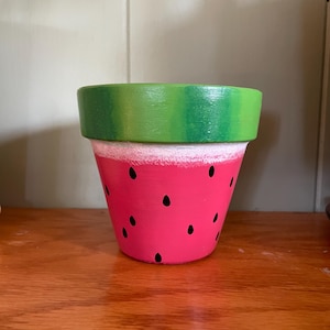 Puede incluir: Una maceta cónica pintada para parecer una sandía. El borde superior es de color verde sólido, seguido de una fina banda blanca y una base de color rosa brillante decorada con semillas negras en forma de lágrima dispersas por la superficie.