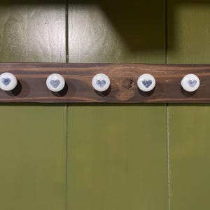 May include: A rectangular dark brown wooden wall rack featuring five white ceramic knobs. Each knob is circular and decorated with a small blue heart pattern. The wooden board has a visible grain and is mounted horizontally with evenly spaced hooks.