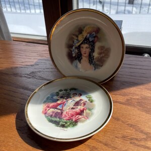 Może przedstawiać: Dwa okrągłe białe talerze porcelanowe ze złotymi obrzeżami. Pionowy talerz przedstawia portret kobiety w żółtym kapeluszu z niebieską wstążką. Przedni talerz ukazuje kolorową scenę z trzema postaciami w czerwono-białych strojach.