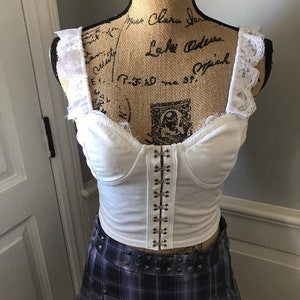 May include: White cropped top with lace trim and a row of silver metal hooks down the front. The top is paired with a purple and black plaid skirt with a studded belt.