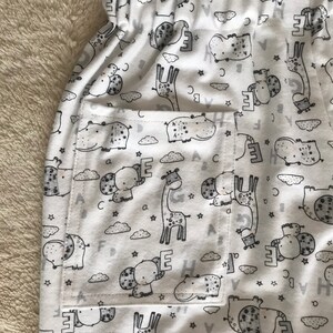 Puede incluir: Tela blanca con un estampado negro y gris de animales y letras. La tela tiene un bolsillo en el lado izquierdo.