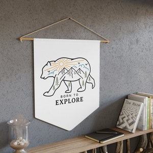 May include: White canvas wall tapestry with a black line drawing of a bear with a mountain landscape inside the bear's silhouette. The sun is shining above the mountains. The text "Born to Explore" is below the bear.