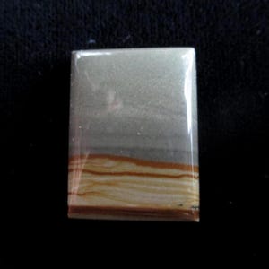 May include: A rectangular gemstone with a polished surface. The top portion is a light gray color, transitioning to a darker gray band, and then to a band of tan and brown horizontal lines. The stone is set against a black background.
