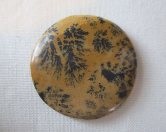 May include: A round, polished stone with a dendritic agate pattern. The stone is a light brown color with black dendritic markings that resemble branches or ferns.