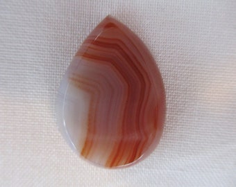 May include: A teardrop-shaped agate cabochon with white and red banding. The stone is polished and has a smooth surface.