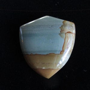 May include: A shield-shaped gemstone with a polished surface. The stone features layers of color, including blue-gray, tan, and brown. The gemstone has a pointed bottom and a rounded top, with a natural, earthy appearance.