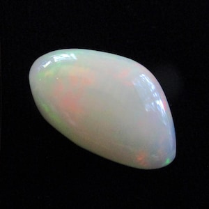 May include: A polished, oval-shaped opal gemstone. The opal has a milky white base color with iridescent flashes of green, pink, and orange. The gemstone is set against a black background, highlighting its luminous quality.