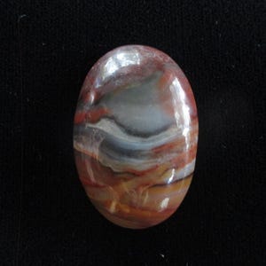 May include: A smooth, oval-shaped cabochon gemstone with a swirling pattern of red, brown, white, and gray colors.