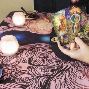 Puede incluir: Una configuraci&oacute;n de lectura de cartas del tarot con velas encendidas, cristales y un mantel decorativo con un dise&ntilde;o floral en negro y rosa. Varias cartas del tarot se sostienen en la mano, mostrando ilustraciones coloridas.