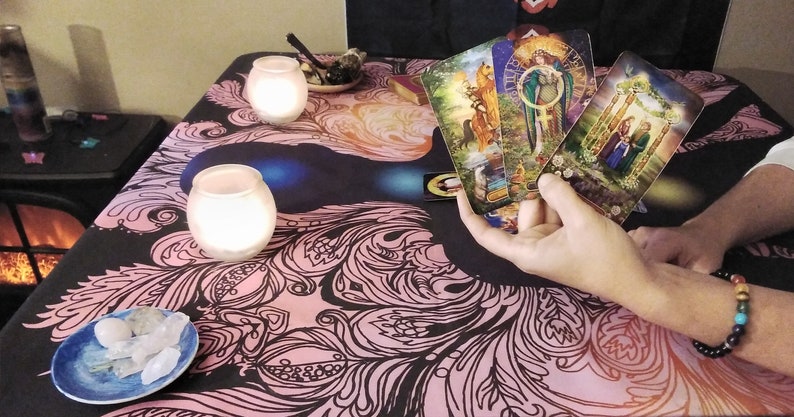 Puede incluir: Una configuraci&oacute;n de lectura de cartas del tarot con velas encendidas, cristales y un mantel decorativo con un dise&ntilde;o floral en negro y rosa. Varias cartas del tarot se sostienen en la mano, mostrando ilustraciones coloridas.