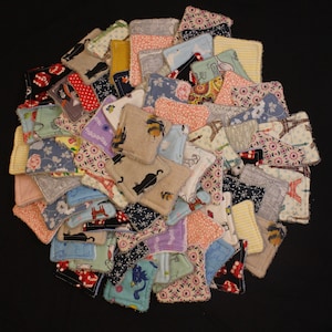 May include: A collection of fabric coasters in a variety of colours and patterns, including floral, cat silhouettes, and geometric designs. The coasters are arranged in a circular shape against a black background. The coasters appear to be handmade.