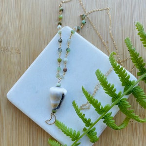 May include: A gold chain necklace featuring a series of small, multi-colored green and brown beads, a single white and brown cone-shaped seashell pendant, and a small gold crescent moon charm dangling at the very bottom of the pendant.