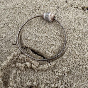 May include: A silver-toned bracelet with a natural seashell accent. The bracelet is circular and rests on a bed of sand. The seashell is a light beige color with darker brown stripes. The bracelet is a piece of jewelry.