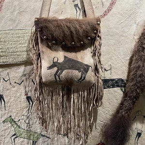May include: A tan suede shoulder bag featuring a dark brown fur flap secured with metal studs. The bag displays a black painted animal graphic on the front and is decorated with long, thin suede fringe along the sides and bottom edges.