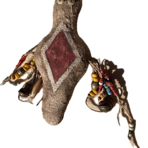 May include: A tan textured fabric doll featuring two small white button eyes, a central red diamond-shaped patch with a white border, and dangling side attachments decorated with multi-colored beads, small metal bells, and a light-colored claw.