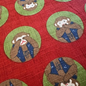 May include: Red fabric with a repeating pattern of green circles containing brown monkeys with blue jackets covering their eyes and mouths. The monkeys are wearing blue jackets with white accents.