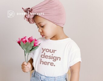 Op de afbeelding: Een jong persoon draagt een wit T-shirt met de tekst "your design here." erop gedrukt. Ze dragen een roze tulband en houden een boeket roze bloemen vast.