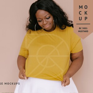 May include: A woman wearing a mustard yellow t-shirt with a white skirt. The t-shirt is a mockup with the text "MOCKUP" and "BC 3001 MUSTARD" on a brown tag.