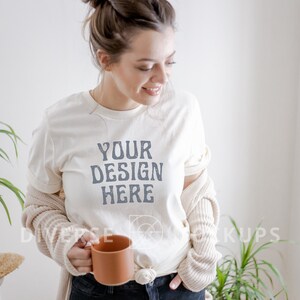 Puede incluir: Una mujer lleva una camiseta blanca con el texto "YOUR DESIGN HERE" impreso en ella. Est&aacute; sosteniendo una taza de cer&aacute;mica marr&oacute;n en su mano izquierda. Lleva una chaqueta de punto beige y pantalones vaqueros azules.
