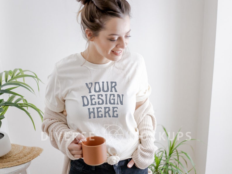 Puede incluir: Una mujer lleva una camiseta blanca con el texto "YOUR DESIGN HERE" impreso en ella. Est&aacute; sosteniendo una taza de cer&aacute;mica marr&oacute;n en su mano izquierda. Lleva una chaqueta de punto beige y pantalones vaqueros azules.