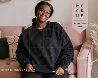 Op de afbeelding: Een zwarte Gildan 18000 sweatshirt mockup op een vrouw die op een roze bank zit. De tekst "MOCKUP" en "GILDAN 18000 BLACK" is zichtbaar in de afbeelding.