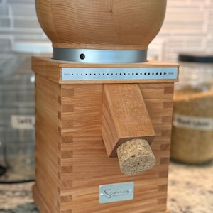 May include: A wooden grain mill with a light brown finish. The mill has a bowl-shaped top, a rectangular body, and a spout with a cork stopper. The words "Fein" and "Grob" are visible on a metal strip. The brand name "Sano" is also visible.