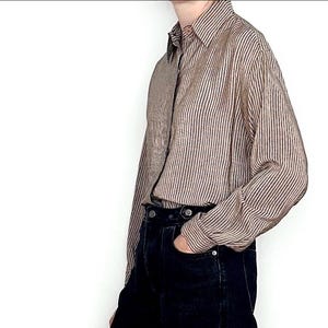 May include: A long-sleeved button-down shirt featuring a vertical pinstripe pattern in shades of brown and cream. The shirt has a classic collar and is tucked into a pair of solid black denim jeans with visible metal buttons at the waistline.