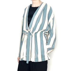 Peut inclure: Haut à manches longues de style cache-cœur à rayures verticales bleu clair et blanc cassé. Il est noué à la taille et porté avec un pantalon noir. Le haut est confectionné dans un tissu doux et fluide.