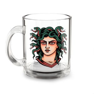 Puede incluir: Una taza de cristal transparente con una colorida ilustración gráfica de Medusa que tiene serpientes de color verde oscuro como cabello, ojos rojos y una expresión facial severa. El diseño está impreso en el frente del recipiente de cristal.