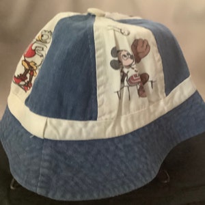 May include: A blue and white bucket hat with a white brim and a white band around the crown. The hat has a patchwork design with images of Mickey Mouse, Donald Duck, and Goofy.