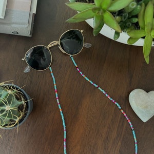 Könnte beinhalten: Eine Sonnenbrille mit einer Perlenkette. Die Kette ist türkis, pink und orange. Die Sonnenbrille ist schwarz und gold.