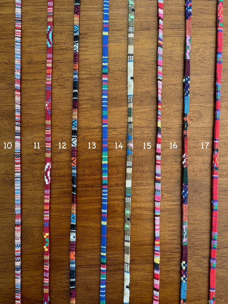 May include: Ten different colorful woven cords with various patterns. The cords are all approximately the same length and are displayed against a wooden background.