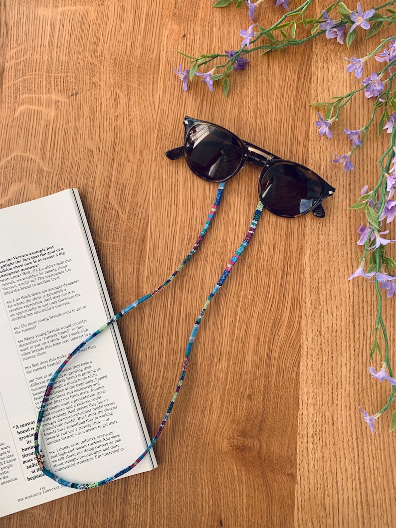 May include: A pair of brown sunglasses with a colorful patterned strap. The strap is made of woven fabric and has a variety of colors, including blue, green, red, and yellow. The sunglasses are lying on a wooden surface with a book and purple flowers.