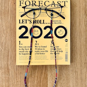 May include: A yellow magazine with the title "The Forecast" and the year "2020" printed in black. The magazine is held by a pair of black glasses with a colorful beaded strap.