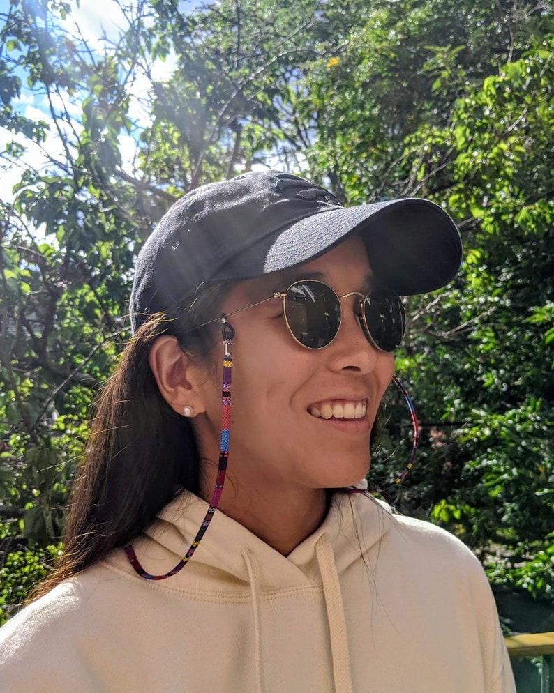 May include: A person wearing a black baseball cap, round sunglasses with a colorful beaded strap, and a cream colored hoodie.